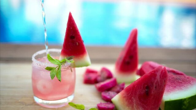 Pouring mineral water into mojito cocktail with red watermelon by the pool