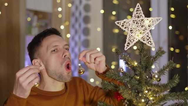 Man takes glass Christmas ball out of mouth and covers face with hand in disgust of situation, shame. Guy protests against festive mood. Concept of denial of holidays, melancholic person, depression.