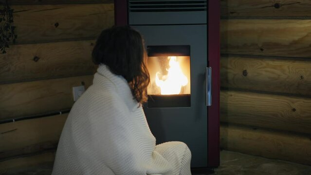 A young Caucasian woman is sitting by an electric pellet fireplace wrapped in a blanket.