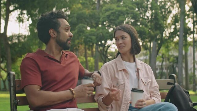 Medium Shot Of Young Man And Woman Sitting On Bench In Park, Having Coffee And Discussing Something While Meeting Outdoors On Summer Day