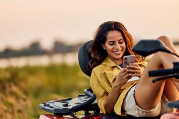 A cheerful woman lying on a quad bike and typing on her mobile phone.