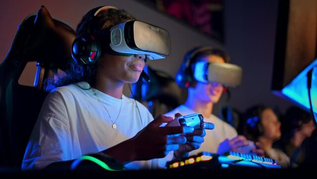 White boy and black girl teens in VR headsets playing video games in video game club with blue illumination using a gamepad