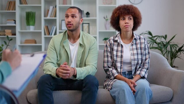 Multicultural married couple visiting psychologist to solve family issues. Young man sharing own vision of problems while woman sitting silently and nervously rubbing hands. Marital therapy concept.