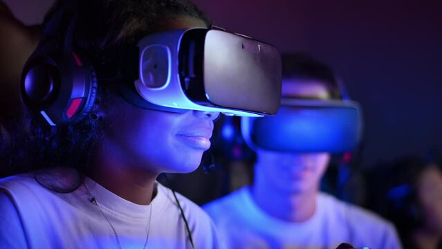 White boy and black girl teens in VR headsets playing video games in video game club with illumination using a gamepad, talking in voice chat. Slow motion