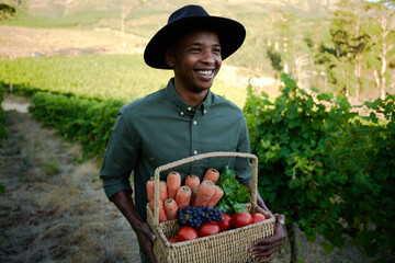 Happy young black man in casual clothing holding basket with fruit and vegetables on farm