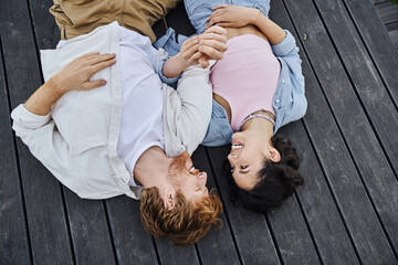 joyful interracial couple lying down on wooden porch and looking at each other outdoors, top view