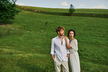 just married interracial couple standing together in green field, scenic and tranquil landscape