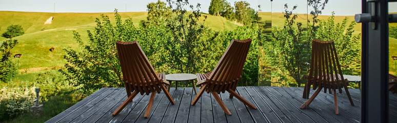 wooden chairs and coffee table on porch with view on scenic landscape near glass building, banner