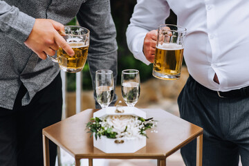 Two male friend drinking beer from glass mugs at a wedding party.