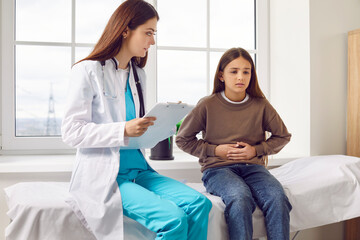 Teenage girl sitting on the couch in the doctor's office and pointing to her stomach to paediatrician during medical examination in clinic. Child doctor listening to the patient's complaints.