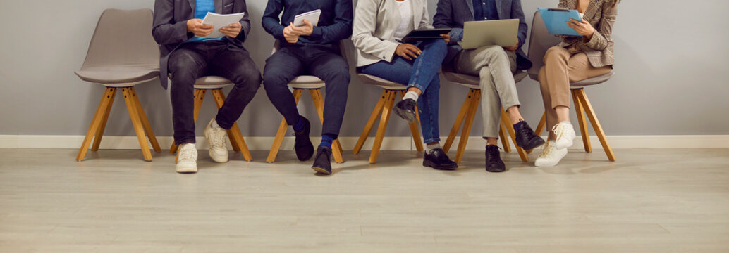 Legs of various people sitting in row in waiting room before interview or business meeting. Pople in smart and casual clothes sit on chairs reading their resumes. Low section. Panoramic web banner.