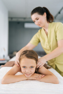Vertical portrait of happy cute little girl having neck, shoulder and back massage by unrecognizable female masseuse at medical clinic, smiling looking at camera. Adorable preteen kid feeling relax.