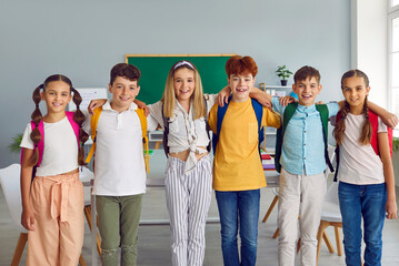 Group of elementary school students standing together in row and hugging. Portrait of happy friends, classmates wearing casual clothes posing in classroom looking at camera. School education concept