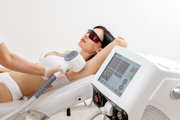 Body care concept. Laser hair removal. Young Caucasian woman in safety glasses lies on the sofa, female cosmetologist doing laser hair removal in a beauty salon - obrazy, fototapety, plakaty