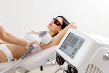 Fotobehang Schoonheidssalon Body care concept. Laser hair removal. Young Caucasian woman in safety glasses lies on the sofa, female cosmetologist doing laser hair removal in a beauty salon