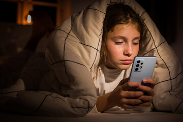 Ten-year-old Caucasian girl at night under a duvet in her room holding a phone looking at the screen almost falling asleep. Sleeping girl cannot tear herself away from games or the news feed. - obrazy, fototapety, plakaty