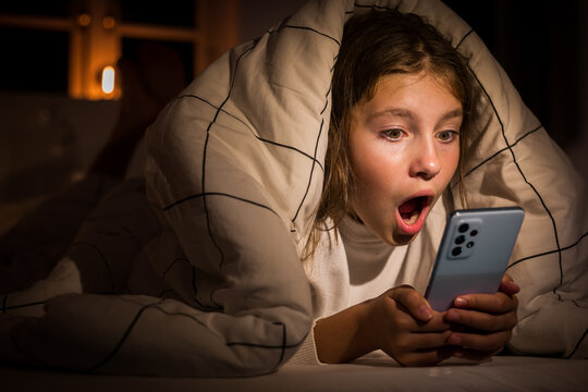 Portrait of surprised shocking face of ten year old Caucasian girl holding looking at phone screen at night lying on the bed under a duvet. Cute girl read a scary story while preparing for Halloween.