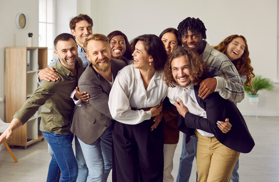 Happy Business Team. Cheerful Diverse Colleagues And Friends Having Fun At Work. Group Of Different Young Smiling Multiracial People In Smart Casual Wear Standing In Office And Hugging All Together