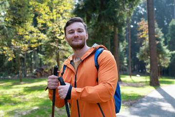 A young healthy man in an orange jacket and a blue backpack in the forest. Smiling man holding trekking poles in his hands