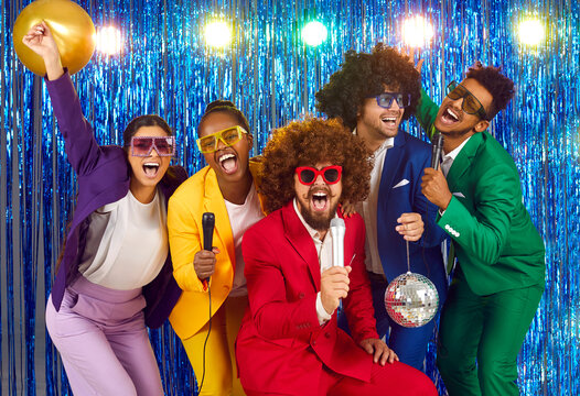 Portrait multinational funny group laughing happy cheerful people dressed in bright business suits, voluminous curly wigs and large disco glasses with rhinestones, singing into microphones in karaoke.