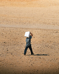 portrait of an Egyptian man walking in the middle of the sandy desert. He is wearing typical Egyptian clothing, with tunic and sandals. He is carrying a jerrycan with water on his back. 