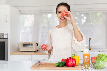 Obraz na płótnie Canvas Happy young latin lady having fun and covering eyes with tomato halves, fooling around in kitchen interior, looking and smiling at camera while cooking dinner