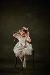 Fototapeta premium Portrait of charming girl, child listening music in headphones sitting on chair against vintage background. Fairy princess or queen. Heroine of story tale.