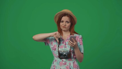 Woman traveller in straw hat holding smartphone and shows thumbs down with negative face expression. Isolated on green background.