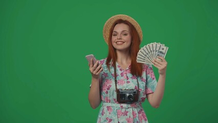 Woman traveller in straw hat holding smartphone and fan of money bills, smiling face expression. Isolated on green background.