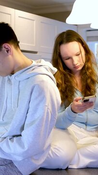 teens sit on table in kitchen girl offended upset pouted boy turned back on phone looks next gadget eat fruit year in background 