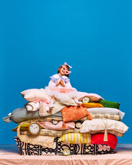 Portrait of little princess in rose sunglasses in heart shape sitting on big pile of pillows over blue background. Fashion fairy character