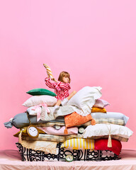 Portrait of angry little princess sitting on big feather bed with pillows with french baguette over pink studio background.