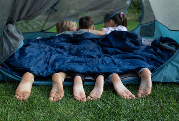 cute bare feet of three children peeking out of sleeping bag. Children, brothers and sisters or friends lie on their stomachs in tent. Healthy lifestyle, little tourist, family activities, summer fun