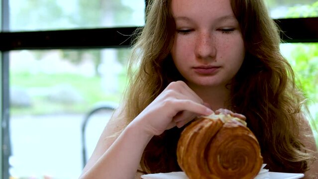 a young girl eats a round croissant in a restaurant by the window, she picks up cream with her little finger and licks the delicious cream from her finger. a delicacy to taste adolescence