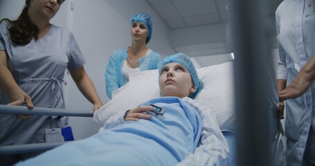 Medics transport patient to hospital room after surgery down medical center hallway. Young girl lies on gurney, looks at pulse oximeter on her finger and talks with mother. Medical staff at work.