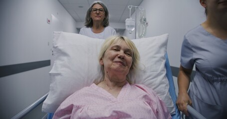 Obraz premium Mature female doctor with young nurse brings senior patient to medical center. Old woman lies on gurney with drip and talks with medics about surgery. Medical staff at work in modern hospital.