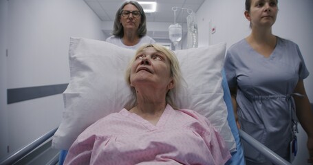 Fototapeta premium Mature female doctor with young nurse brings senior patient to medical center. Old woman lies on gurney with drip and talks with medics about surgery. Medical staff at work in modern hospital.