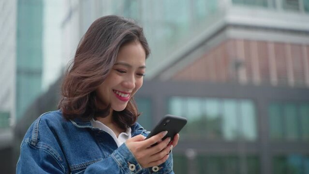 Young asian woman using mobile phone in the urban city outdoor happy pretty girl looking at cellphone smile in front of office building