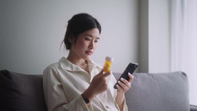 Sick  Asian woman holding a pill bottle reading information medicine label instructions or prescription when get flu on sofa in living room at home, Healthcare and medicine concept.