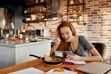 Young happy caucasian woman using a smartphone while having breakfast in the morning