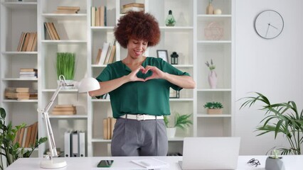 Pretty curly african american woman confessing in love while making heart gesture with hands. Young female office worker showing true feelings and expressing positive emotions at workplace. - Powered by Adobe