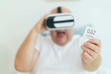 Caucasian oversize fat man showing a condom in his hand while watching an entertainment in the virtual reality glasses or goggles.