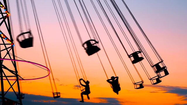 Unrecognizable silhouettes of people ride on the chained carousel against the warm evenning sky at sunset with at amusement park. Entertainment, summer, fun and holiday concept