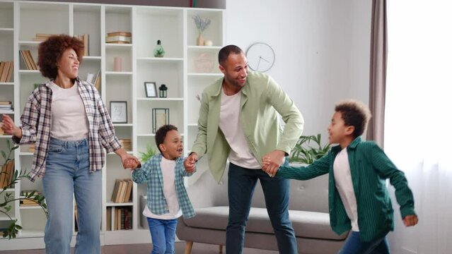 Happy african american family of four spending great time together at home. Joyful parents and two lovely boys dressed in casual attire holding hands and dancing together in spacious living room.