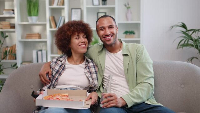 Front view of delightful african american spouses taking time to relax and enjoying comedy movie together. Cheerful man and woman dressed in every-day clothes eating pizza and drinking cold drinks.