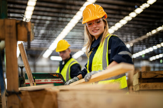 woman cleaning timber wood in dark warehouse industry. Team worker carpenter wearing safety uniform and hard hat working and checking the quality of wooden products at workshop manufacturing. - Powered by Adobe
