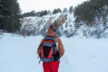 Rear view young man in red brown warm clothes with backpack walking in snow among rocks and cliffs in winter Active lifestyle hiking