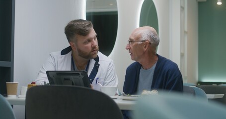 Male doctor shows medical test results to senior patient on digital tablet computer. Adult medic talks with elderly man about treatment in medical center cafeteria. Patient eats meal in hospital cafe.