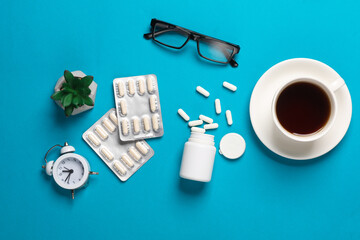 Blisters and bottles of pills, coffee cup, eyeglasses, alarm clock on a blue background. Doctor's workspace. Top view