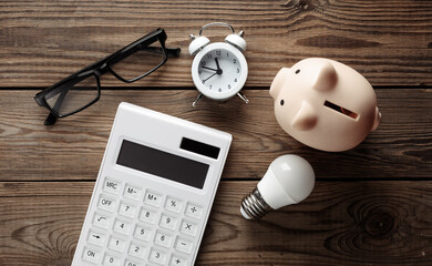 Calculator with eyeglasses , piggy bank, ight bulb and alarm clock on wooden table.  Business concept. Energy saving. Top view - obrazy, fototapety, plakaty